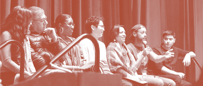 A panel of seven people of multiple races speaking on a stage