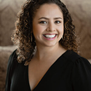Alma Aguilar sits smiling in front of a brown, defocused background in a black v-neck long sleeve shirt.